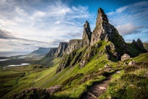 A dramatic view of the Old Man of Storr rock formation on the Isle of Skye, with rugged cliffs, green valleys, and a winding trail.
