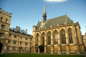 Exeter College Chapel, a grand Gothic chapel with a tall spire, large stained-glass windows, and intricate stone carvings, surrounded by a grassy quad and adjacent historic buildings.