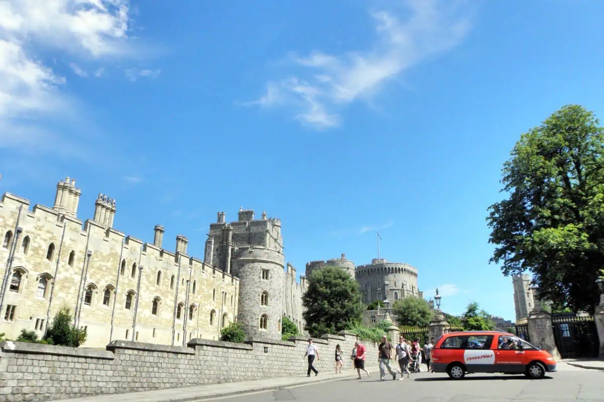 An orange Spaceships Beta Campervan driving past Windsor Castle, with tourists walking along the sidewalk and a clear blue sky.