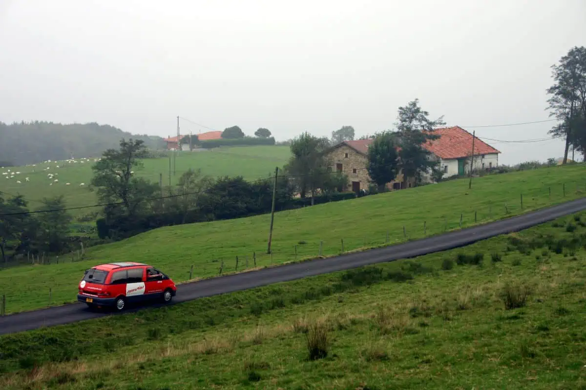 An orange Spaceships Beta Campervan driving along a winding rural road, surrounded by lush green fields, stone farmhouses, and rolling hills under a cloudy sky.