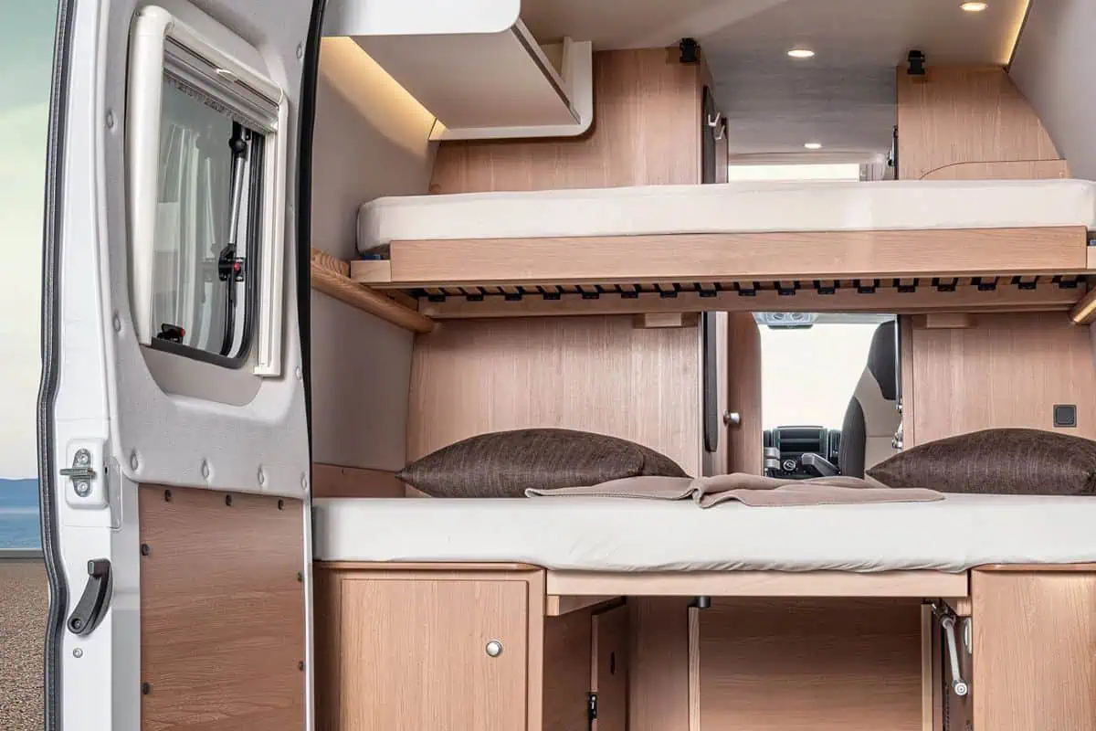 Interior of a camper van with bunk beds, each equipped with pillows and bedding. The lower bunk can be converted into a seating area, and the van features wooden cabinets for storage.