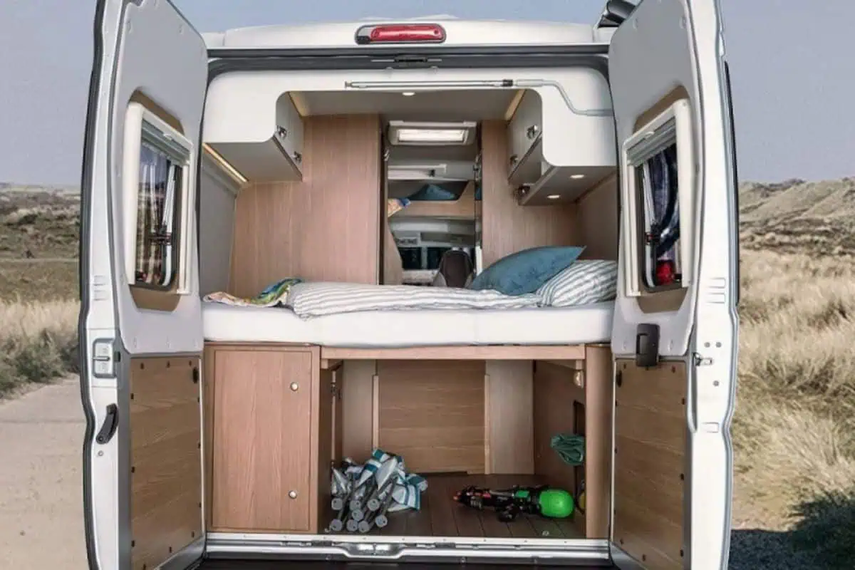 Open rear doors of a camper van revealing a well-organized interior with a raised bed, storage compartments, and a lower storage area containing outdoor gear. The van is parked in a natural setting with dunes in the background.