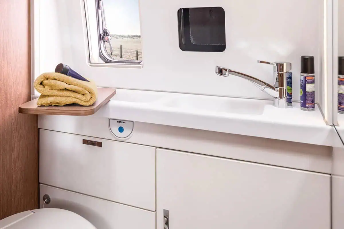 Compact bathroom inside a camper van, featuring a sink, faucet, mirror, and a small window with a beach view. Towels and toiletries are neatly arranged on the counter.