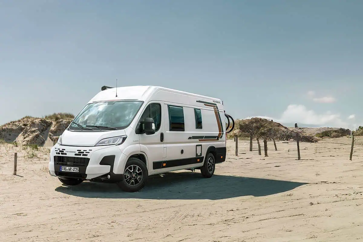 Exterior side view of a white camper van with a sliding door open, parked near a beach. The van features a modern design with black and brown accents.
