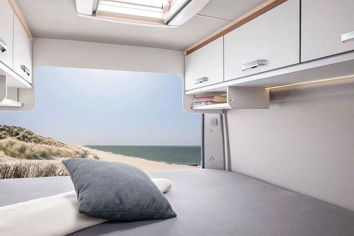 Interior of a camper van showing a cozy sleeping area with a large window offering a panoramic view of a sandy beach and the ocean. A pillow rests on the bed, and overhead cabinets provide storage.
