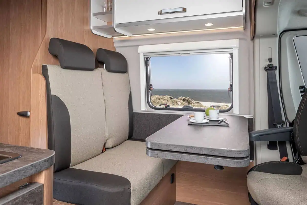 Interior of a camper van showing a seating area with swivel chairs and a foldable table positioned near a window. The table is set with coffee cups and offers a view of the beach and dunes.