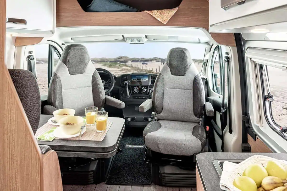 Interior view of a camper van showing the driver and passenger seats swiveled to face a small table with bowls of food and drinks. The van offers a panoramic view of the beach and dunes.