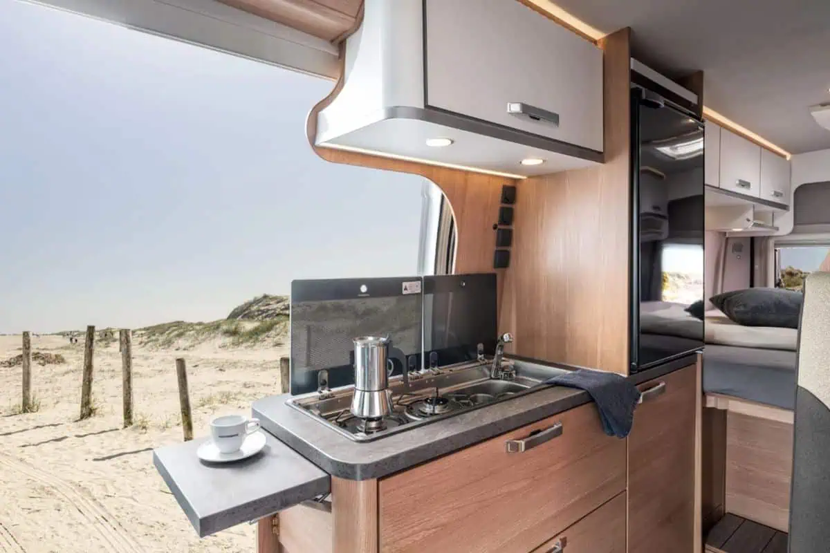Interior of a camper van kitchenette with a gas stove, sink, and overhead cabinets. A coffee cup and coffee maker are placed on the counter, with a beach and dunes visible through the window.