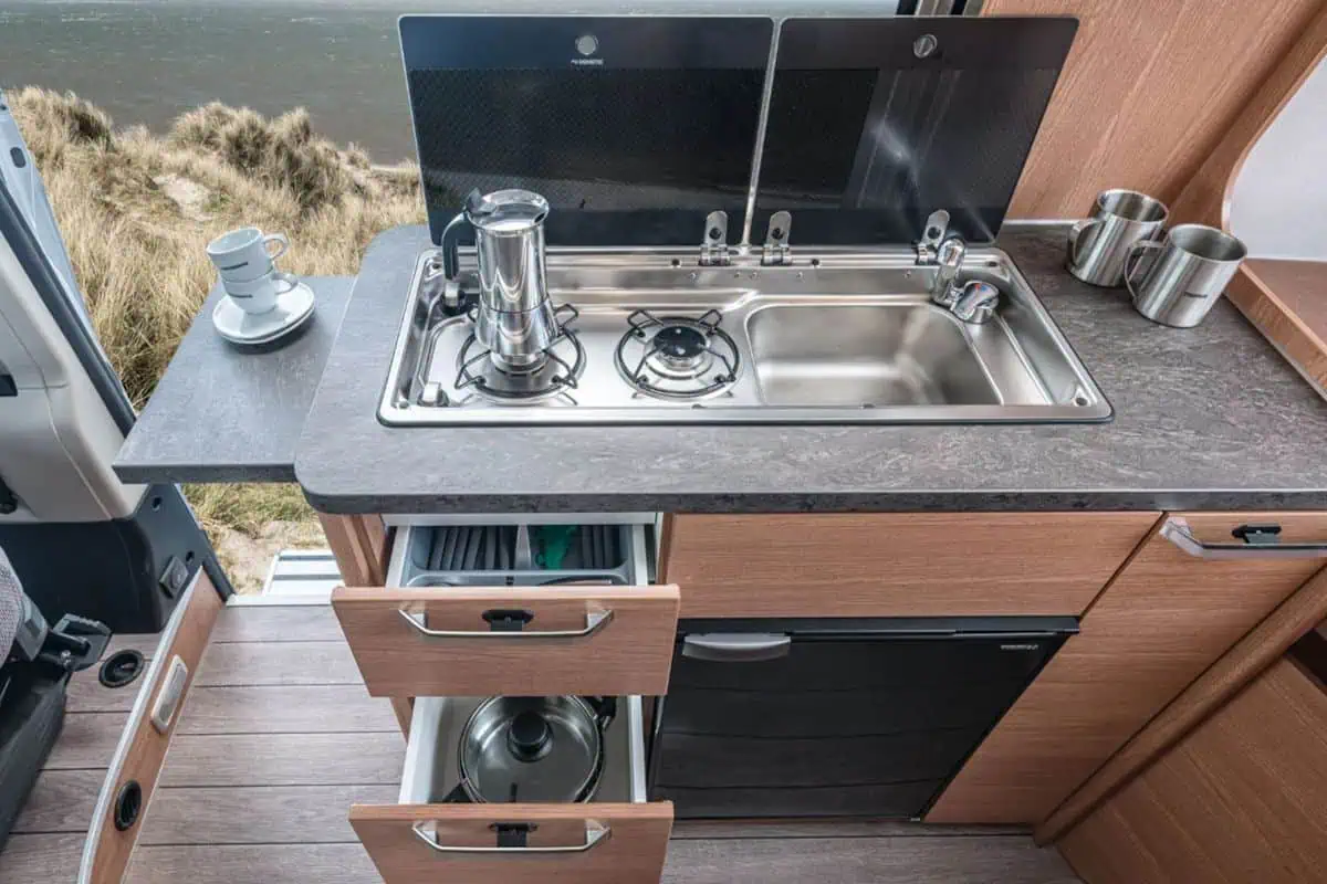 Interior of a camper van showing a compact kitchenette with a gas stove, sink, and storage drawers. A coffee maker and mugs are placed on the countertop, with a scenic view of the beach and dunes outside the window.