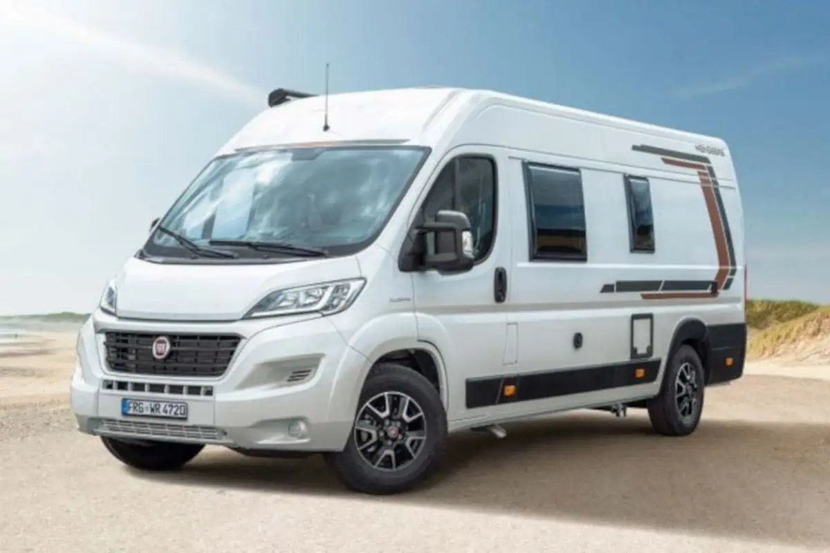Exterior side view of a white camper van with a sliding door open, parked near a beach. The van features a modern design with black and brown accents.