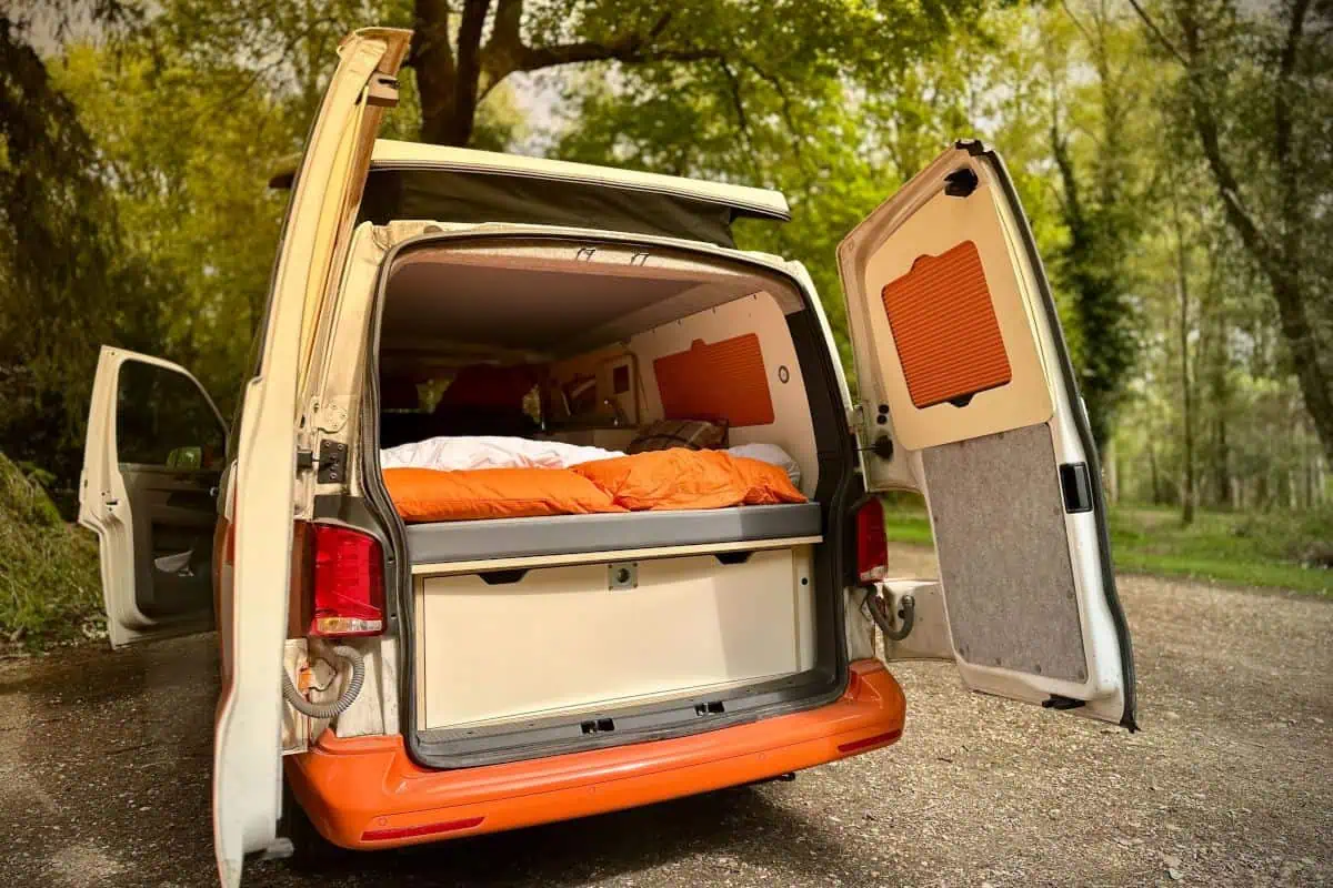 Rear view of an orange and white VW Cosmo Campervan with its trunk and pop-up roof open, showing a bed inside and surrounded by trees.