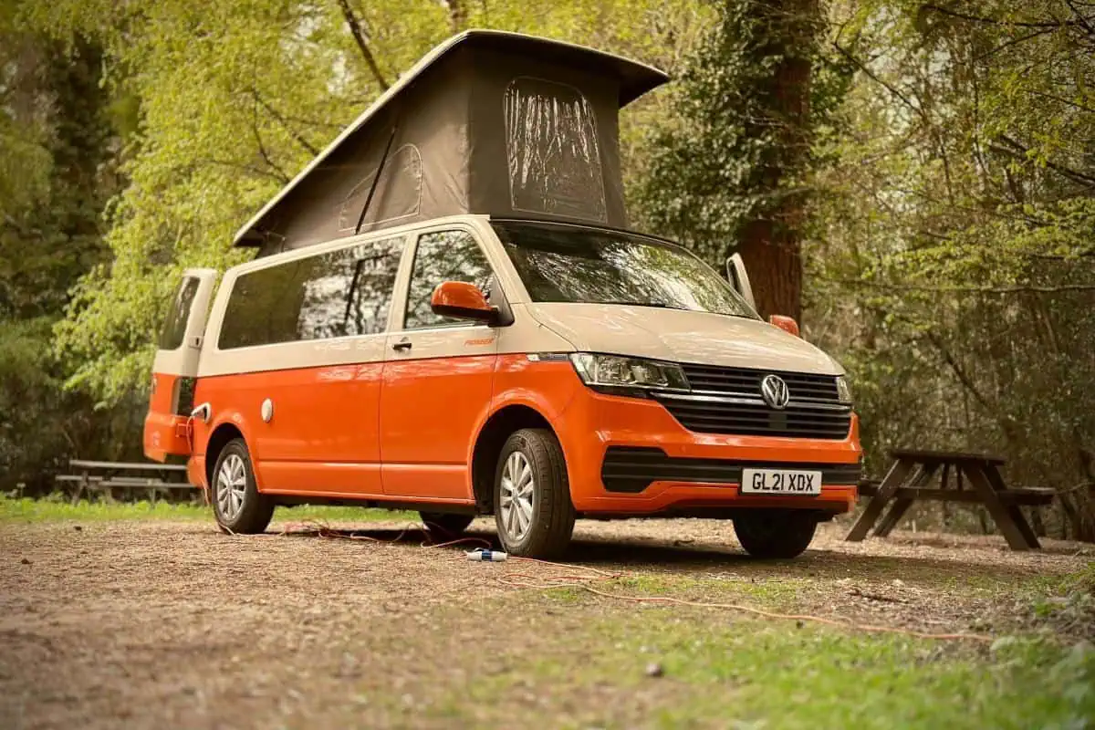 Side view of an orange and white VW Cosmo Campervan parked in a wooded area, with the sliding door and pop-up roof open.