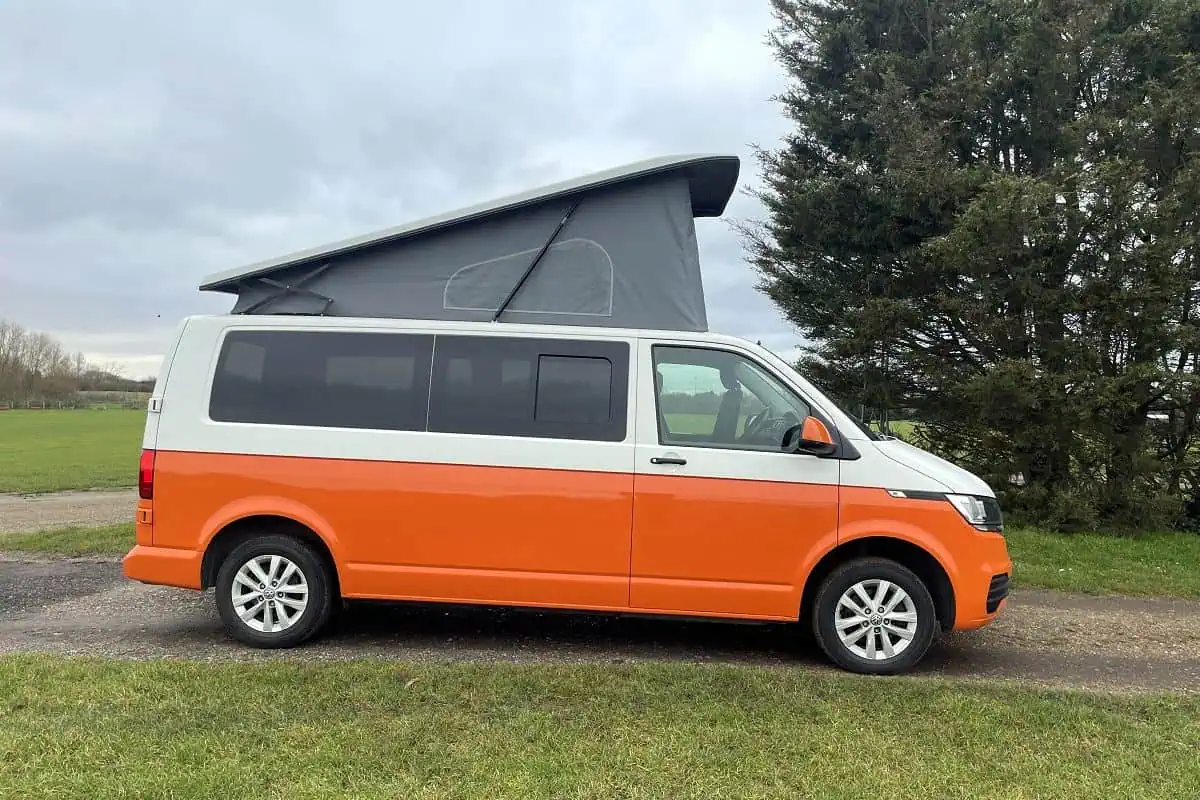Side view of an orange and white VW Cosmo Campervan parked on a gravel path, with a grassy field in the background.
