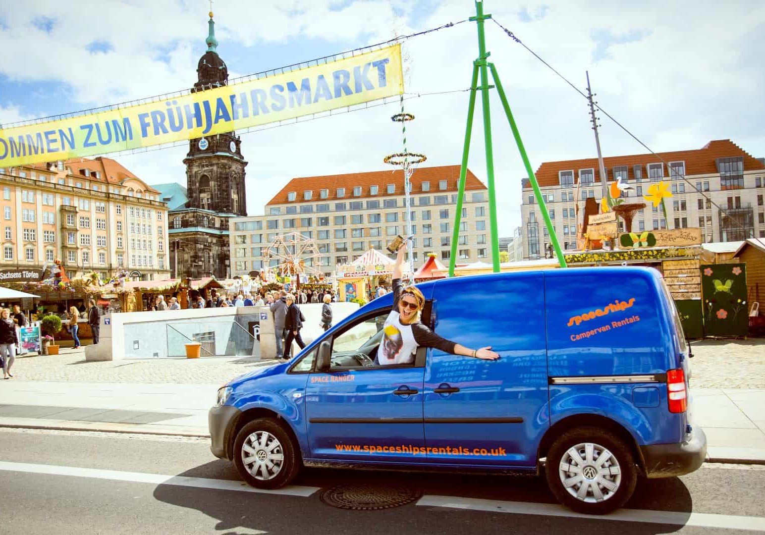 Camper car (blue) parked on German market square with banner with text Spring Fair visible above. Happy Space Traveller hanging out the window of the vehicle.